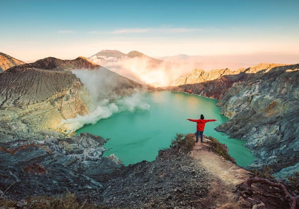 A person stands on a rocky edge with arms outstretched, overlooking a turquoise volcanic lake surrounded by rugged mountains under a clear sky.