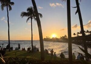 Sunset over the Pacific from the lawn outside The Beach House restaurant on Kauai's South Shore, with palm silhouettes and visitors gathered at the water's edge