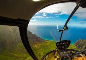 View from a helicopter cockpit flying over green valley ridges toward the ocean on a Kauai helicopter tour
