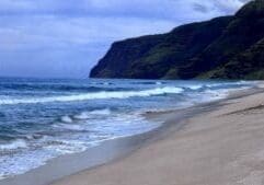 Empty sand beach at Polihale State Park on Kauai with waves rolling ashore and the green cliffs of the Na Pali Coast rising in the distance.